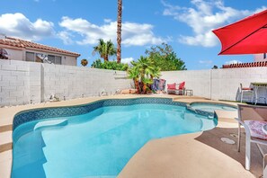 Una piscina con borde de azulejos, una sombrilla roja y un patio con sillas y una mesa.