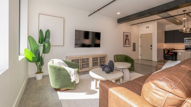 A modern living room with a leather couch, green armchair, and a small round table with decorative items. A large flat-screen TV is mounted on the wall, and there is a potted plant in the corner. - 2BR Stylish Beacon Retreat in Tulsa (Tulsa)