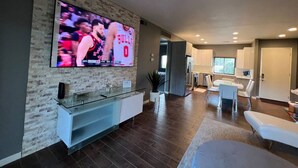 Ein modernes Wohnzimmer mit einem großen Flachbildfernseher an einer strukturierten Wand, einem Glastisch und einer Sitzecke mit weißen Sofas.