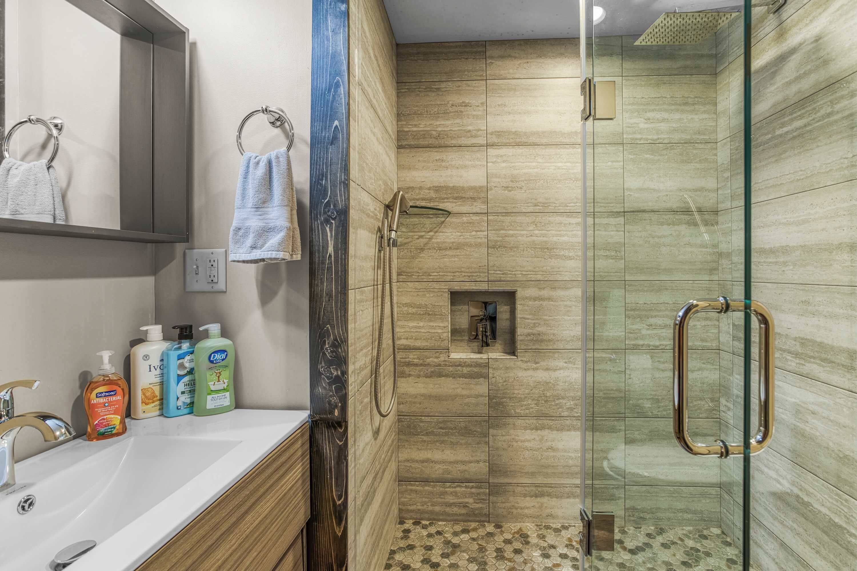 A bathroom with a glass-enclosed shower, a vanity with a mirror, and toiletries on the counter.