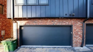 Een moderne garage met een donkere deur, een bakstenen gevel en een groene afvalbak.