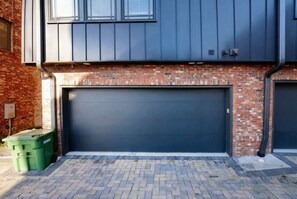 Eine moderne Garage mit dunkler Tür, Ziegelaußenwand und einem grünen Müllbehälter.