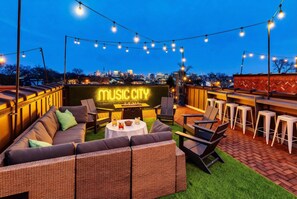 Un área de estar al aire libre con un letrero de neón que dice “MUSIC CITY”, mobiliario de mimbre e iluminación con guirnaldas.