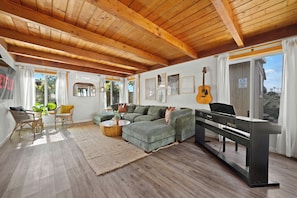 Un salón espacioso con techo de madera, un sofá seccional verde, un piano y un amplio ventanal con cortinas.