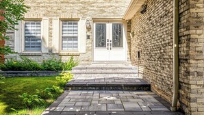 Un bâtiment en briques avec une porte et des fenêtres blanches, un petit jardin devant et un chemin pavé menant à l’entrée.