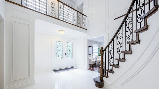 Un hall d’entrée spacieux doté d’un sol blanc poli, d’un escalier à double révolution avec des rampes en métal ouvragé et d’une porte en verre ornée de motifs décoratifs.