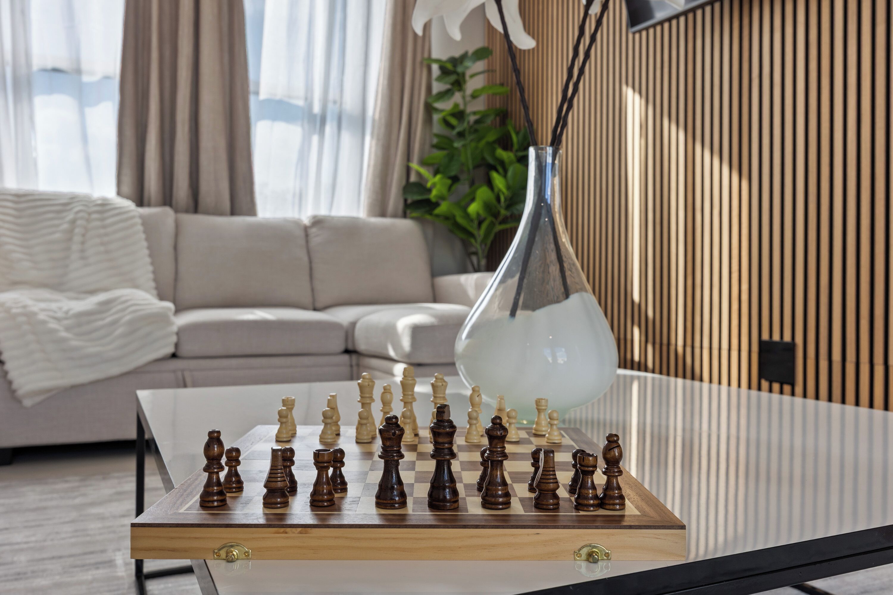 A chessboard on a glass table in a modern living room with a sofa and a plant in the background.