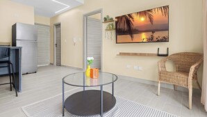 A modern living room with a glass coffee table, a wicker chair, a flat-screen TV on the wall, and a refrigerator in the background. - Cozy 1 BR Apartment by Vistacana (Punta Cana)