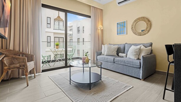 A modern living room with a grey sofa, a wicker chair, a glass coffee table, and a view of a building through large windows. - Cozy 1 BR Apartment by Vistacana (Punta Cana)