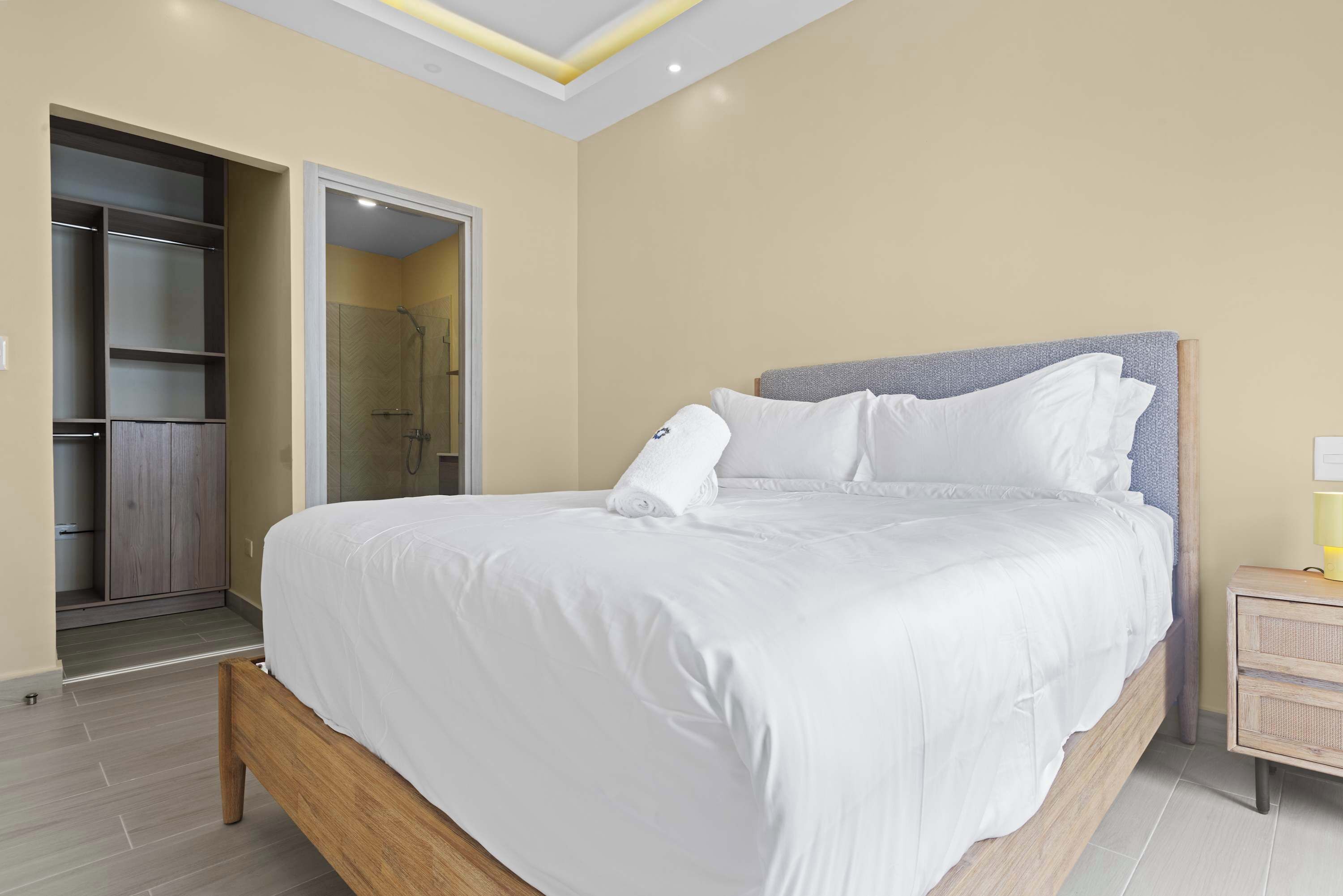 Un lit bien fait, avec du linge de lit blanc, dans une chambre moderne dotée d’une tête de lit en bois, d’un dressing et d’une petite table de chevet avec une lampe.