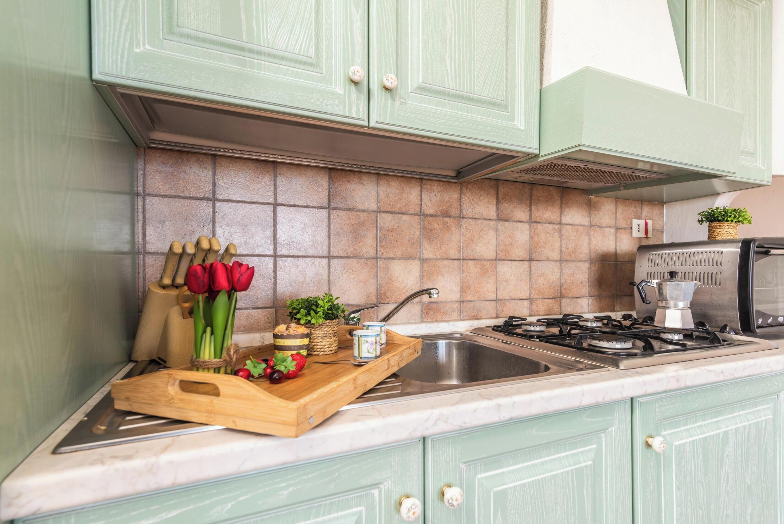 A kitchen with green cabinets, a tiled backsplash, a wooden cutting board with vegetables, and a gas stove.