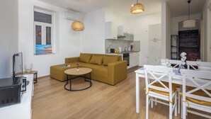 A modern living room with a sofa, a dining table with chairs, a kitchen area, and a large window with a painting. - Anna | 4-Bedroom City Apartment (Rijeka)
