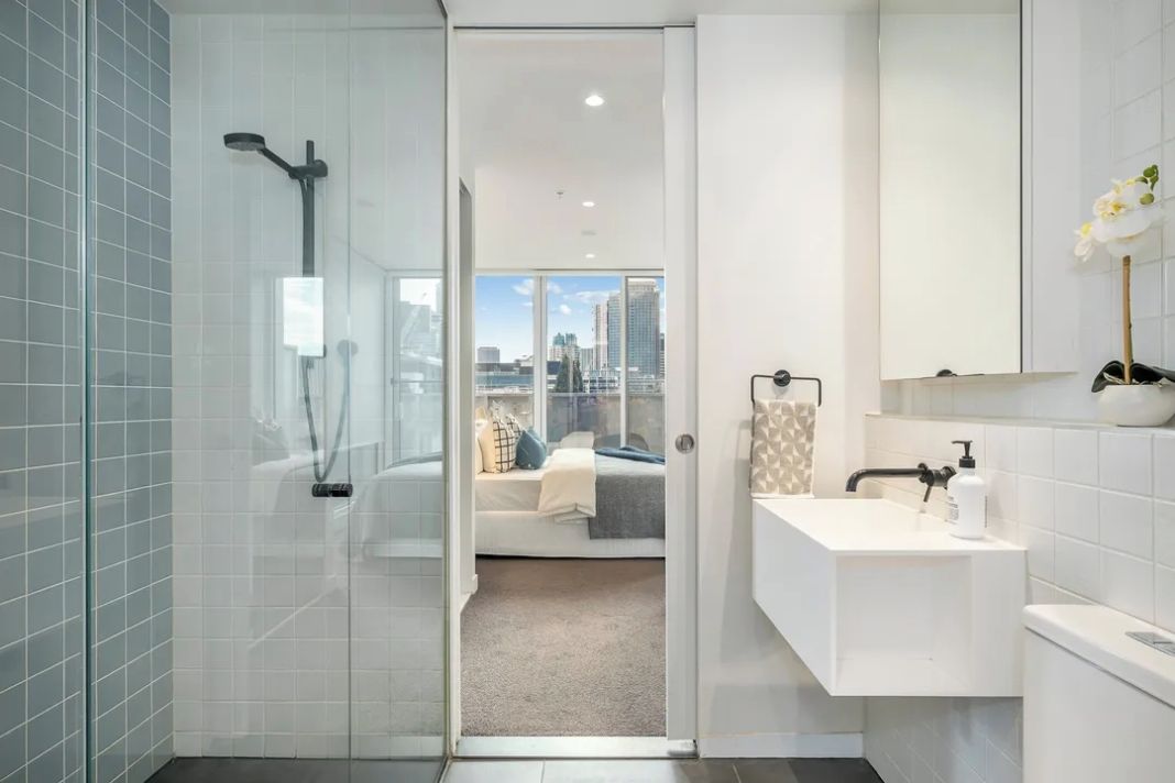 A modern bathroom with a glass shower enclosure, a white sink with a mirror, and a view of a cityscape through an open door.