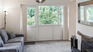 A modern living room with a grey sofa, a hanging pendant light, a fireplace, and a view of greenery through the windows. - 3 bedroom accommodation in Cinderford (Cinderford)