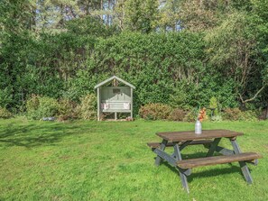 Una mesa de picnic con bancos y una pequeña estructura de madera en un área cubierta de césped, con follaje denso al fondo.