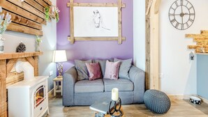 Een gezellige woonkamer met een grijze bank, een houten wand met een decoratie van een hertenhoofd, een schilderij van een paard in een lijst, een klok en een kleine witte verwarming.