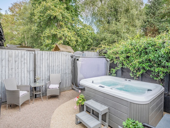Un jacuzzi, sillas de mimbre y una mesita en un patio trasero con cerca de madera y follaje.