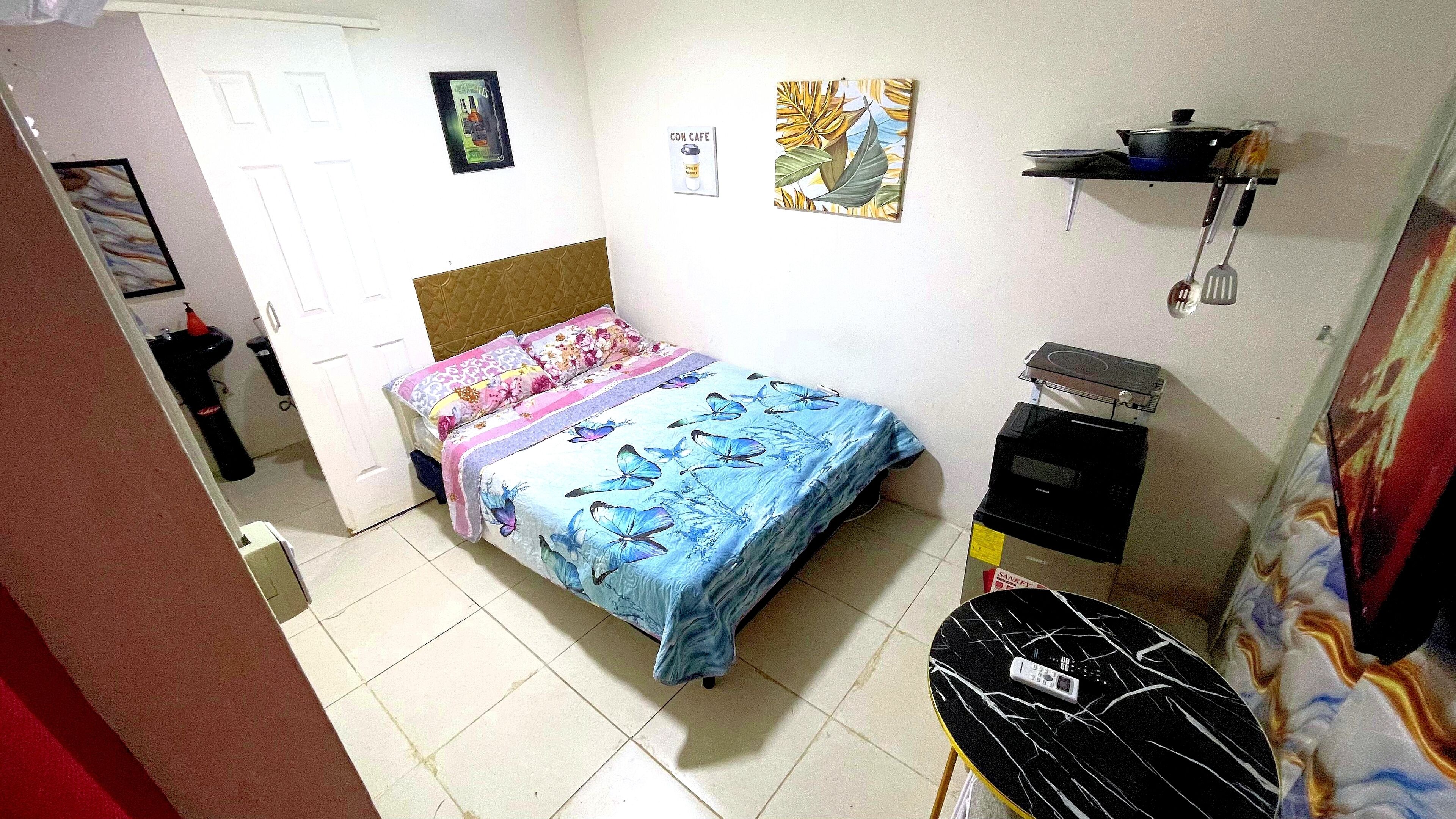 A bedroom with a bed, a small table, a shelf with kitchen items, and a wall with two pictures.