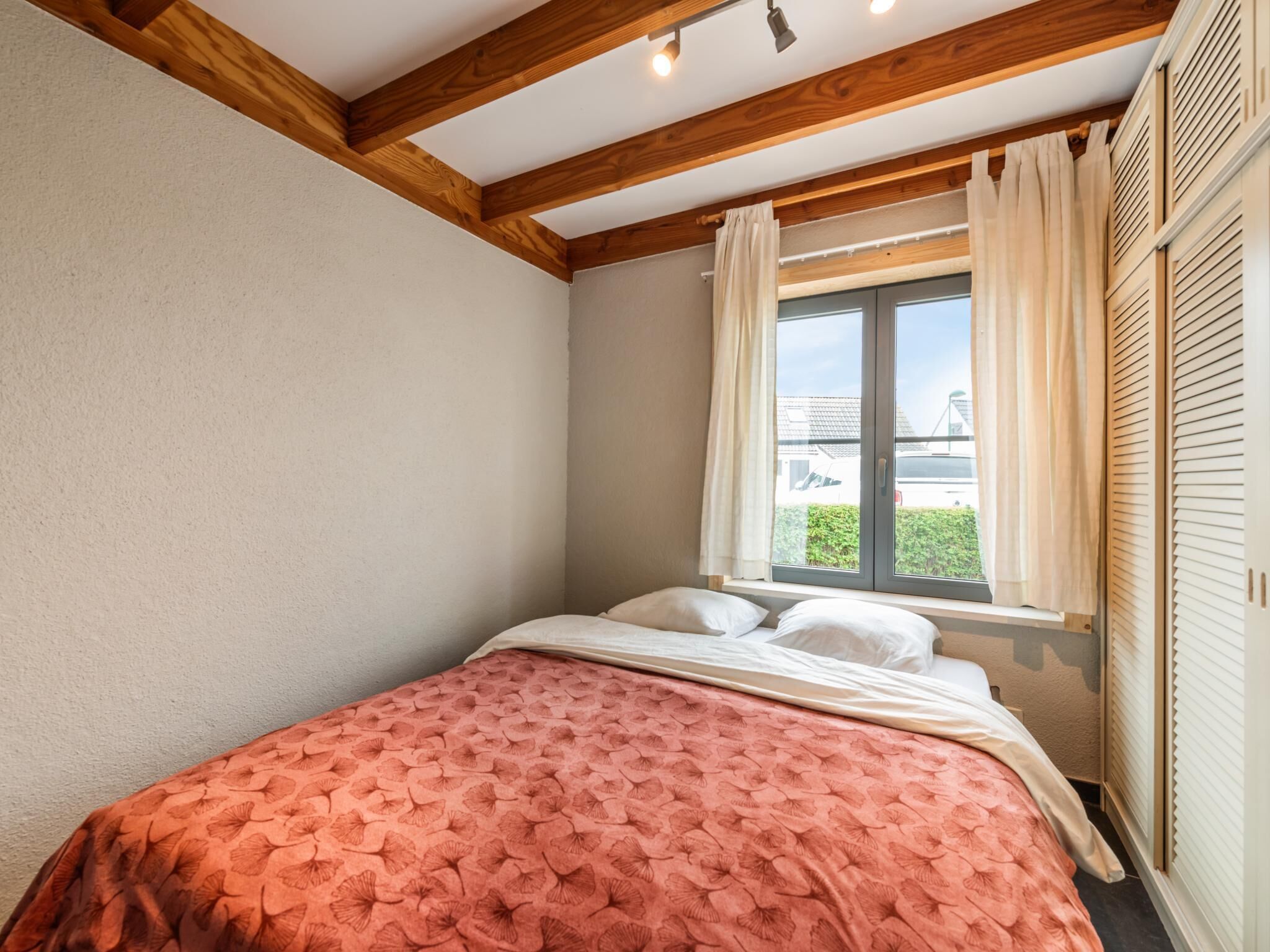 A bedroom with a bed, a window with curtains, wooden beams on the ceiling, and a view of the outside.