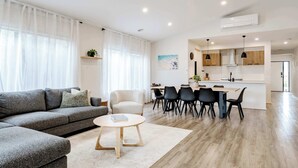 A modern living room with a sofa, coffee table, and dining area with chairs and a kitchen in the background. - Coastal Family Retreat - Relaxed Living with Spa (Torquay)