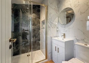 Un baño con cabina de ducha de vidrio, pared con mosaicos de mármol, lavabo con grifo dorado y espejo redondo.