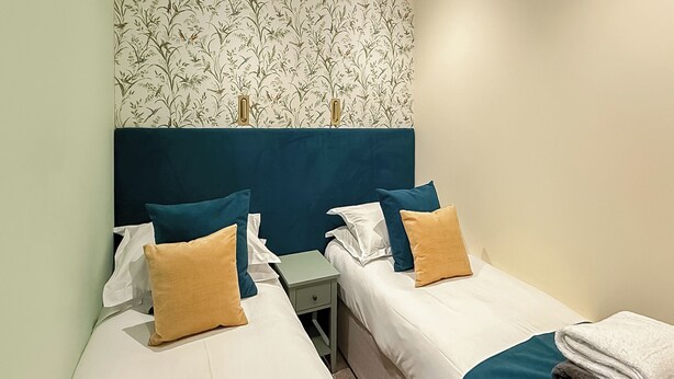 A hotel room with two single beds, a nightstand, a small table, and a floral wallpaper. - 1 bedroom accommodation in Millom (Millom)