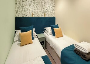 Habitación de hotel con dos camas individuales, una mesita de noche, una pequeña mesa y papel tapiz con motivos florales.