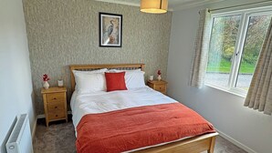 A bedroom with a wooden bed, white bedding, a red throw, a nightstand with a vase, a framed picture on the wall, and a window with curtains. - 1 bedroom accommodation in Llanon (Llanon)