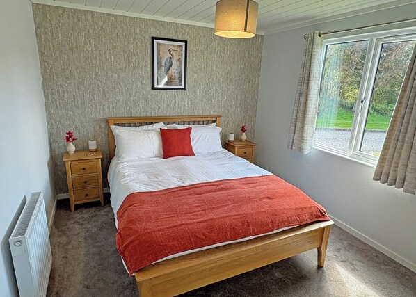 Un dormitorio con una cama de madera, ropa de cama blanca, un plaid rojo, una mesita de noche con un florero, un cuadro enmarcado en la pared y una ventana con cortinas.