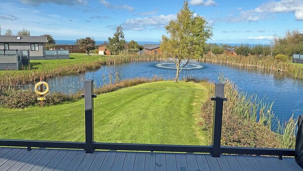 A lakeside view with a grassy area, a wooden deck, and a clear sky. - 1 bedroom accommodation in Llanon (Llanon)