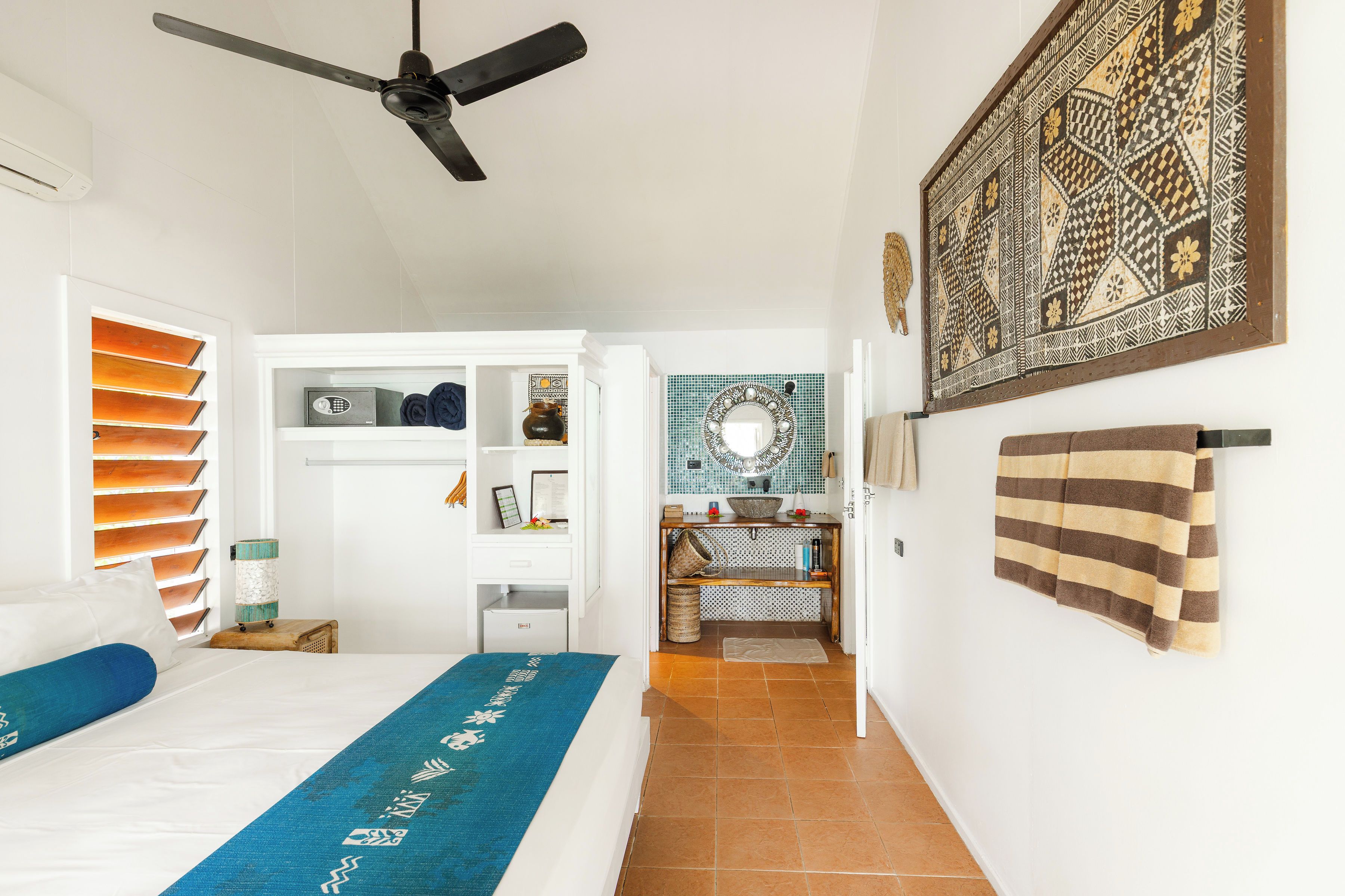 A bedroom with a bed, a ceiling fan, a wardrobe, a bathroom with a sink, and a decorative wall piece.