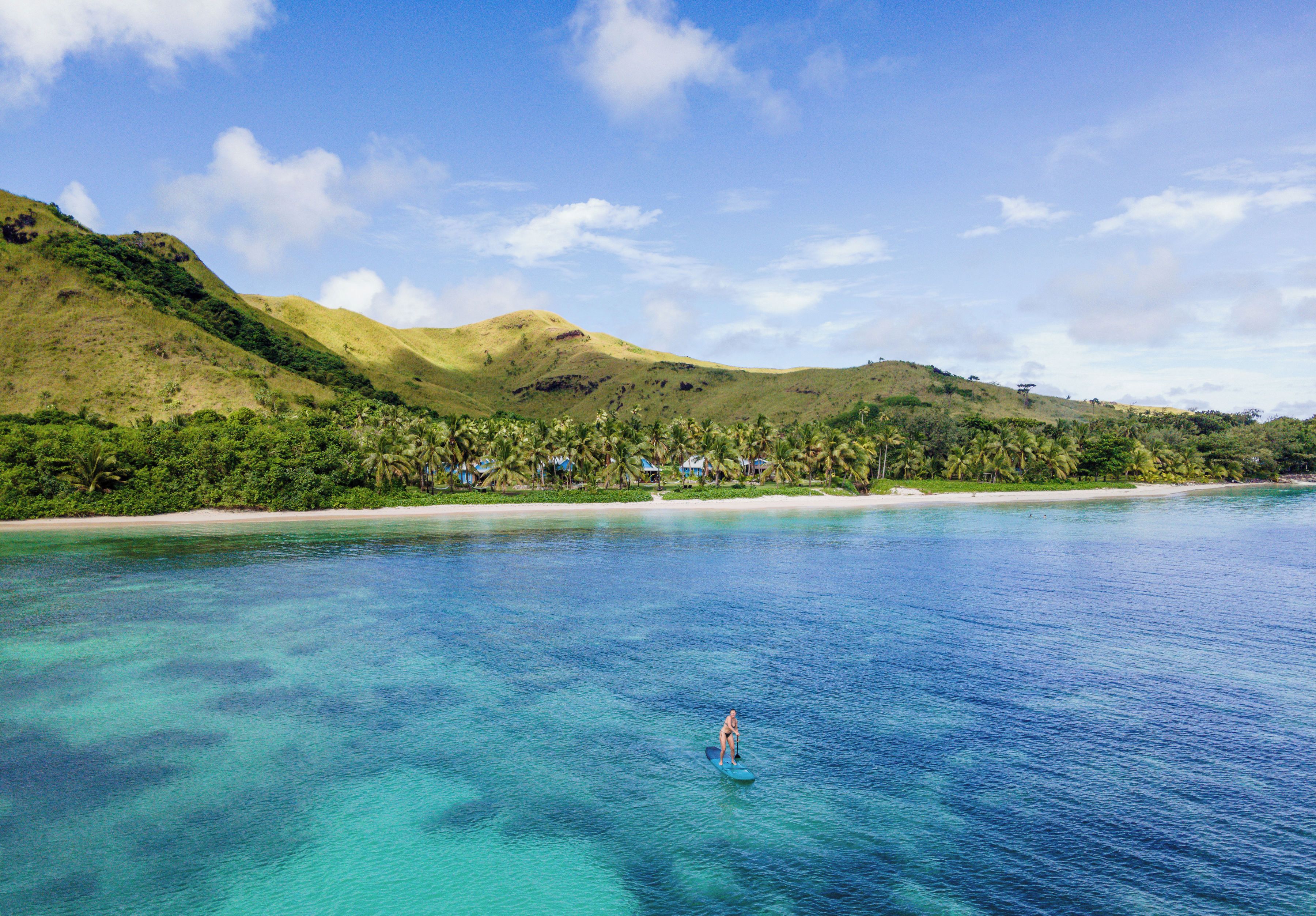 Ein Strand mit klarem türkisblauem Wasser, eine Person beim Paddleboarding, mit sanften grünen Hügeln und Palmen im Hintergrund.