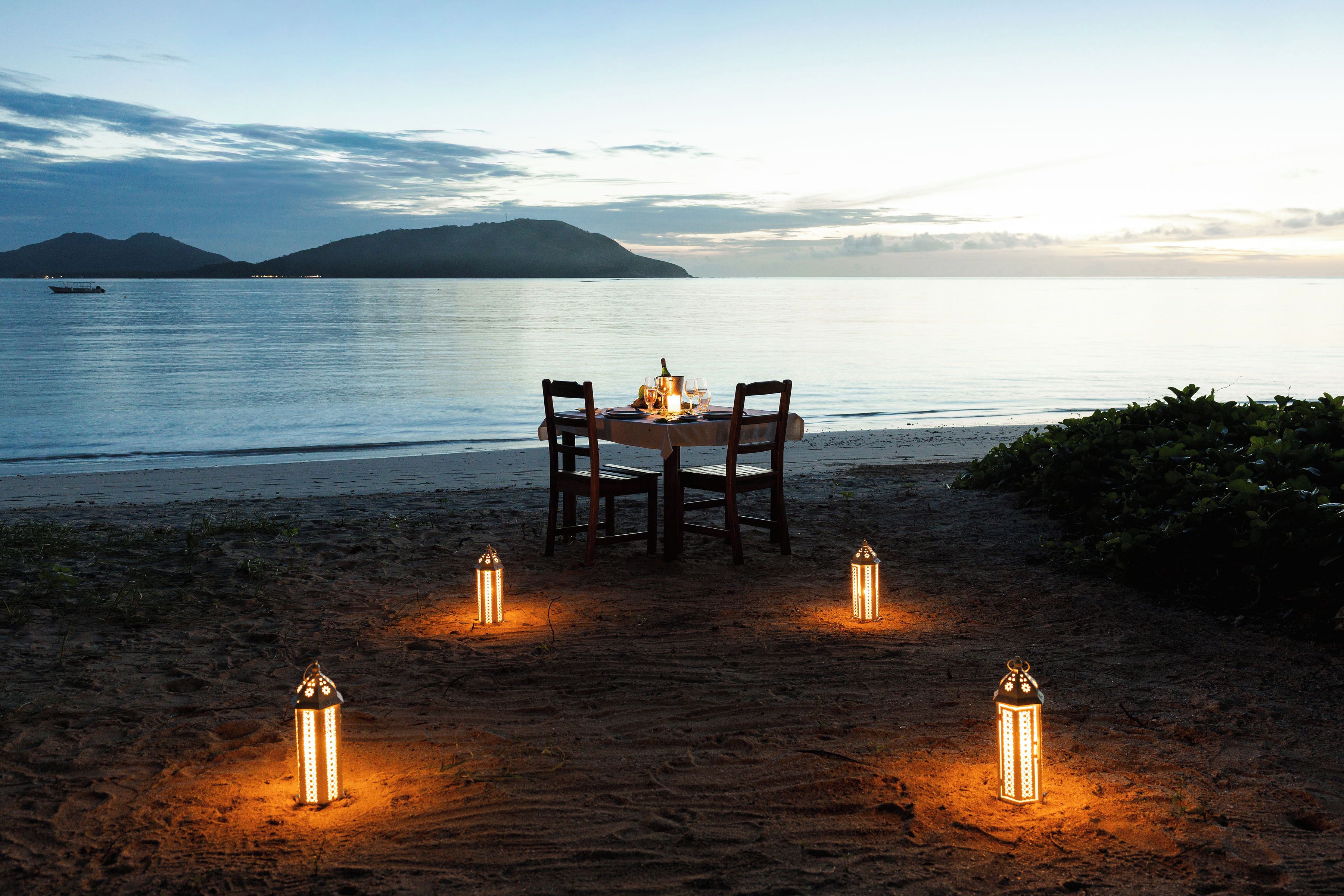 A table set for two on a sandy beach with a view of the sea and a distant island.