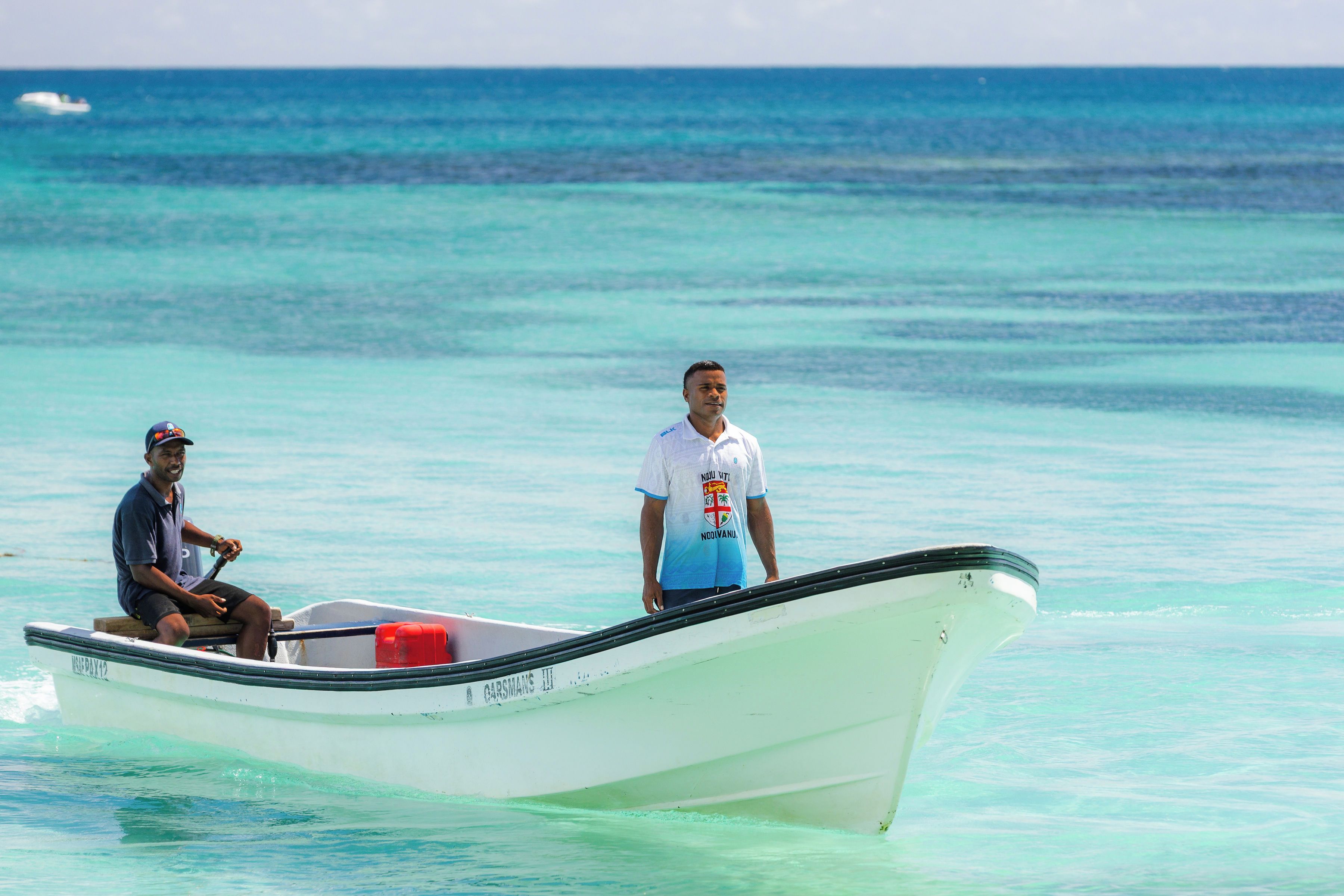 A small boat with two people on it, one seated and one standing, in clear turquoise waters.