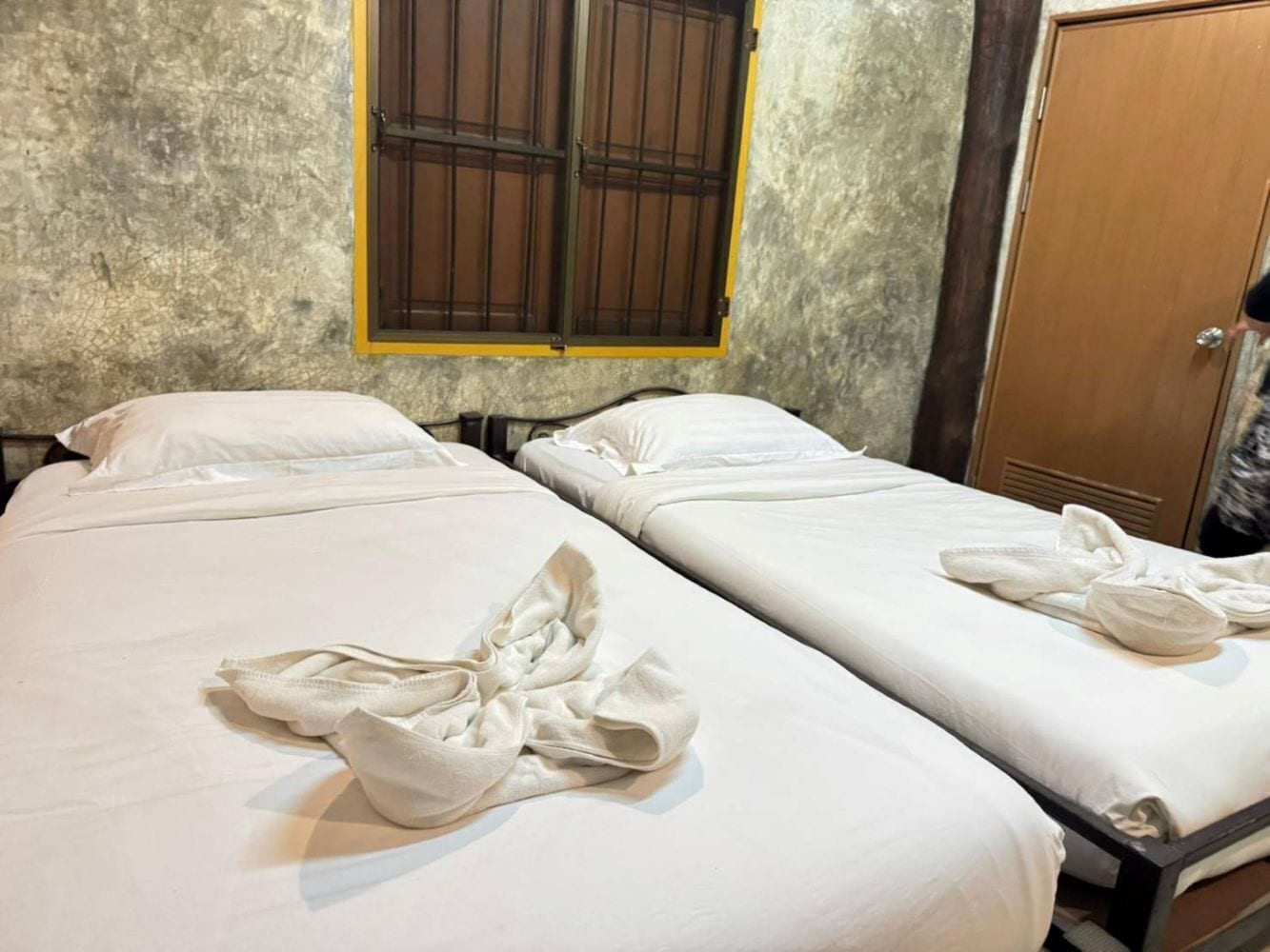 Two single beds with white linens and towels, a window with wooden bars, and a wooden door.
