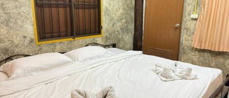 Una cama doble con sábanas y toallas blancas, una puerta de madera y una ventana con reja de metal.