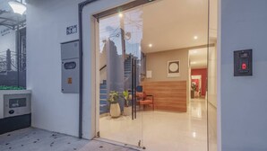 Egy modern szállodahol, üvegajtóval, egy felsőbb szintre vezető lépcsővel, valamint egy fogadópulttal, melyhez egy fa asztal és székek tartoznak.
