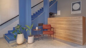 Uma área de recepção moderna com um balcão de madeira, uma cadeira vermelha, duas plantas em vasos, uma mesa redonda pequena e uma escadaria com degraus azuis.