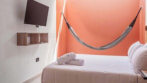 Quarto de hotel com uma cama, uma TV fixada na parede, uma rede e uma toalha com a inscrição 'NOK'.