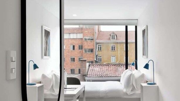 A modern hotel room with a bed, bedside tables, a desk, and a window offering a view of buildings. - NOK Modern Apartment in Chapinero (Bogotá)