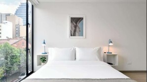 A modern bedroom with a large bed, two bedside tables, a framed picture on the wall, and a view of buildings through a large window. - NOK Modern Apartment in Chapinero (Bogotá)