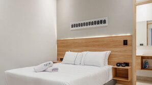 Une chambre d’hôtel moderne avec une tête de lit en bois, un lit bien fait avec du linge de lit blanc, et une petite table de chevet sur laquelle se trouvent une horloge et une télécommande.