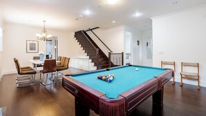 A pool table in a well-lit room with a dining area and a staircase.