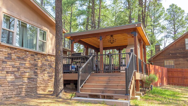 A wooden deck with a staircase leading to a covered area, surrounded by trees. - Royal Heaven (Ruidoso)