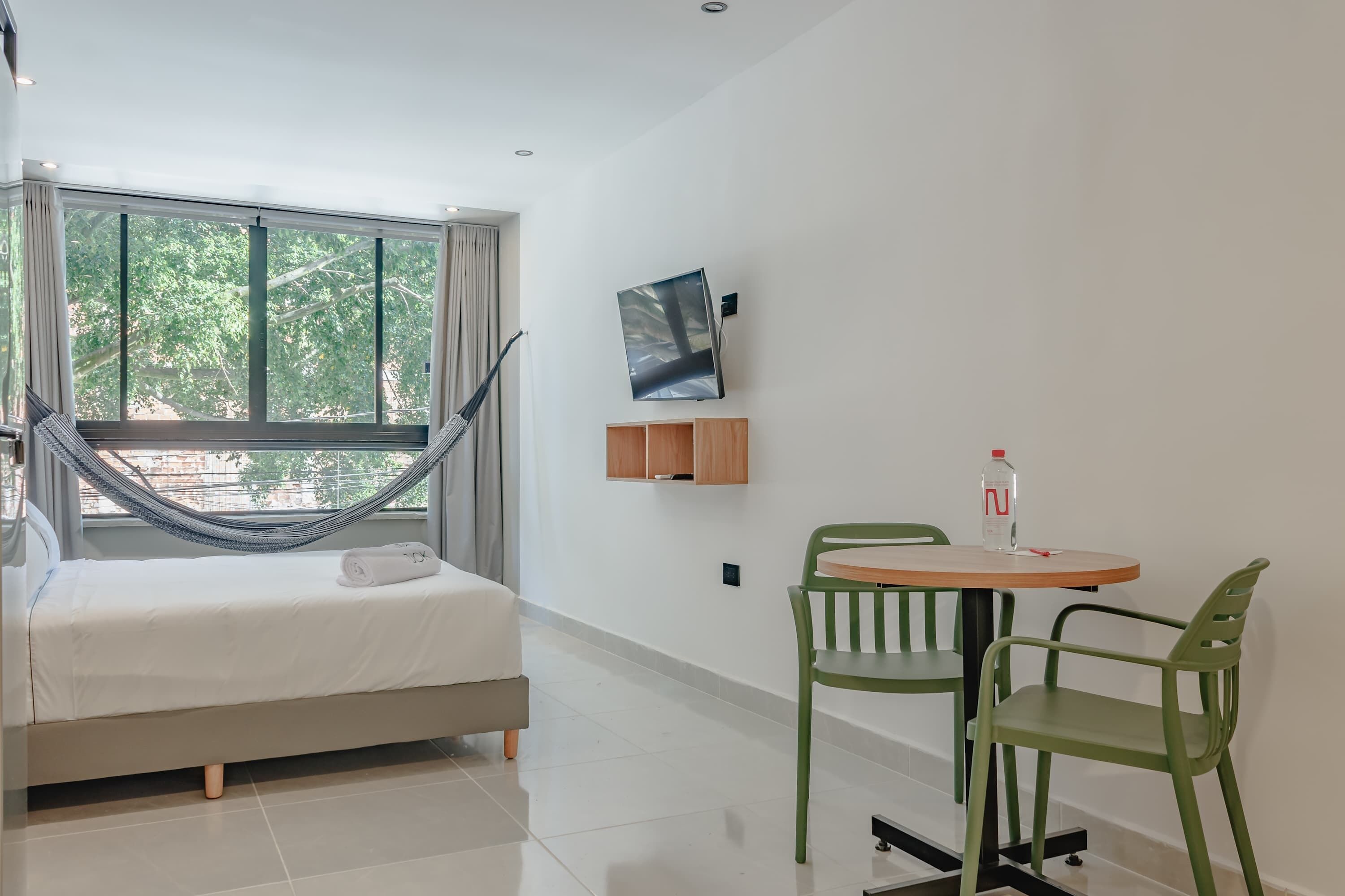 A modern hotel room with a bed, a hammock, a small table with chairs, and a large window offering a view of trees.