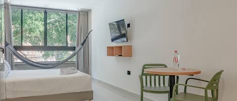 A modern hotel room with a bed, a hammock, a small table with chairs, and a large window offering a view of trees.