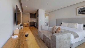Une chambre d’hôtel moderne avec un grand lit, un canapé, une table à manger en bois avec un verre à vin, et un coin cuisine à l’arrière-plan.