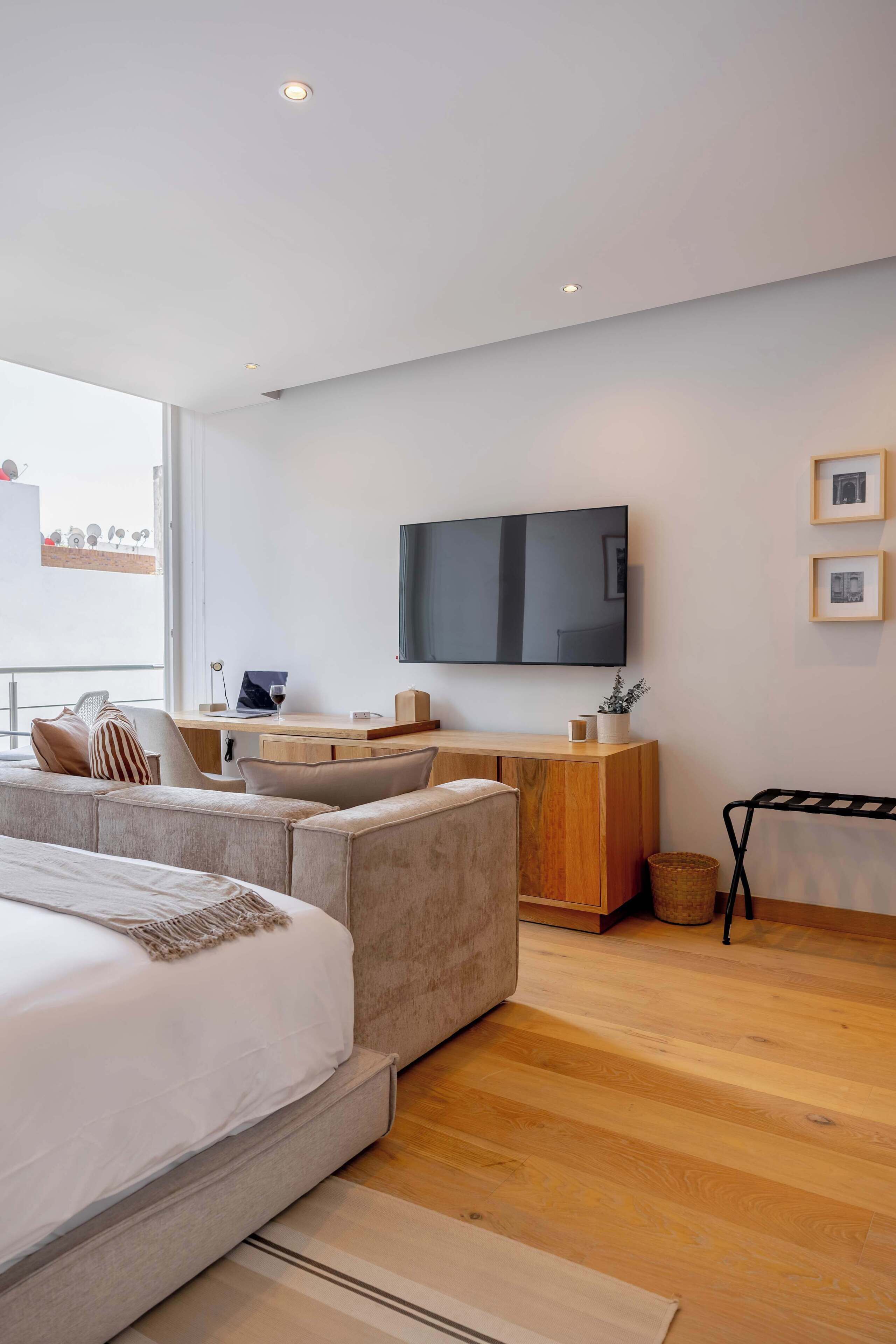 A modern hotel room with a large bed, a sofa, a desk with a laptop, and a TV mounted on the wall.