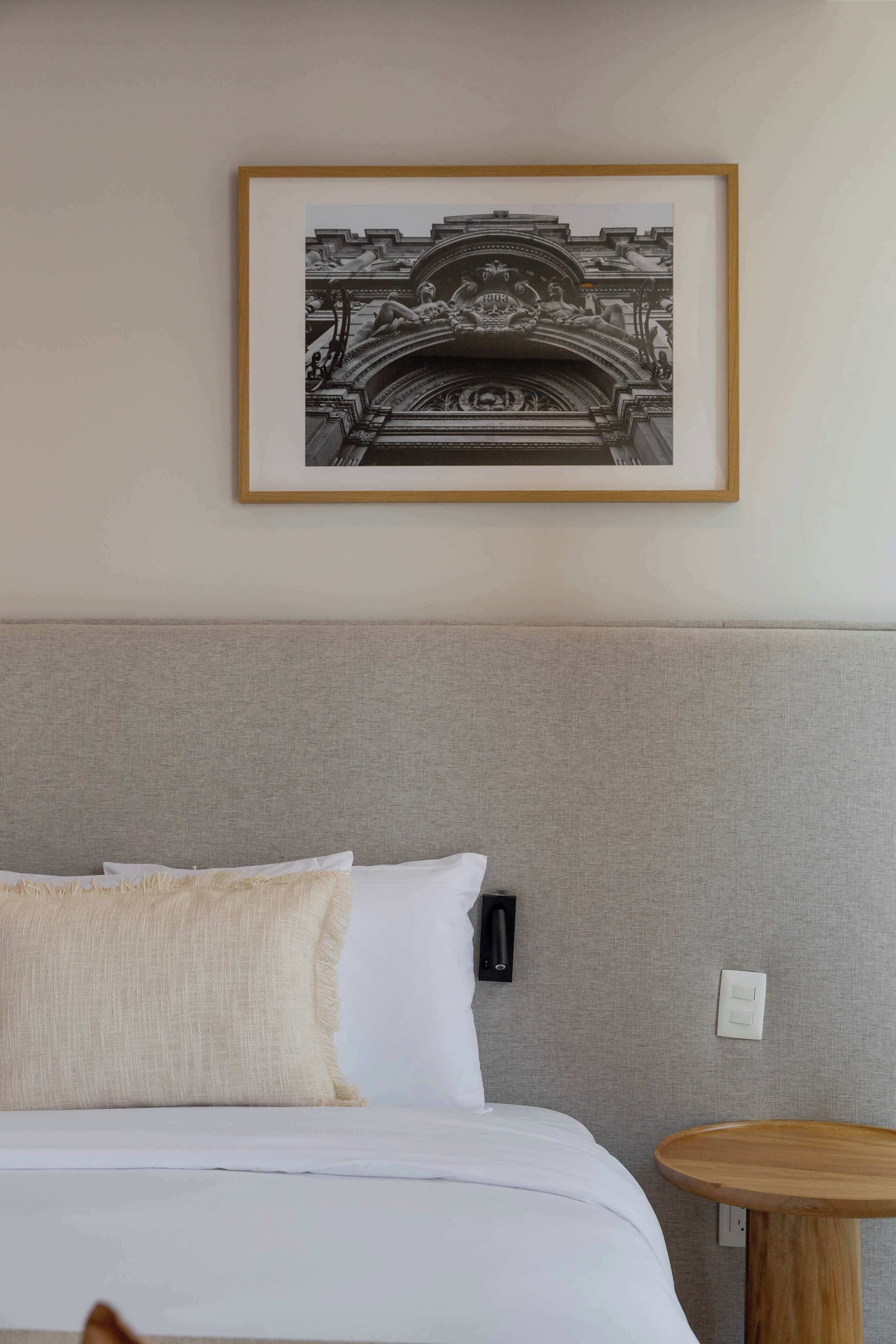 Quarto de hotel com uma cama, uma mesa lateral redonda de madeira, uma fotografia em preto e branco emoldurada na parede e uma cabeceira cinza texturizada.