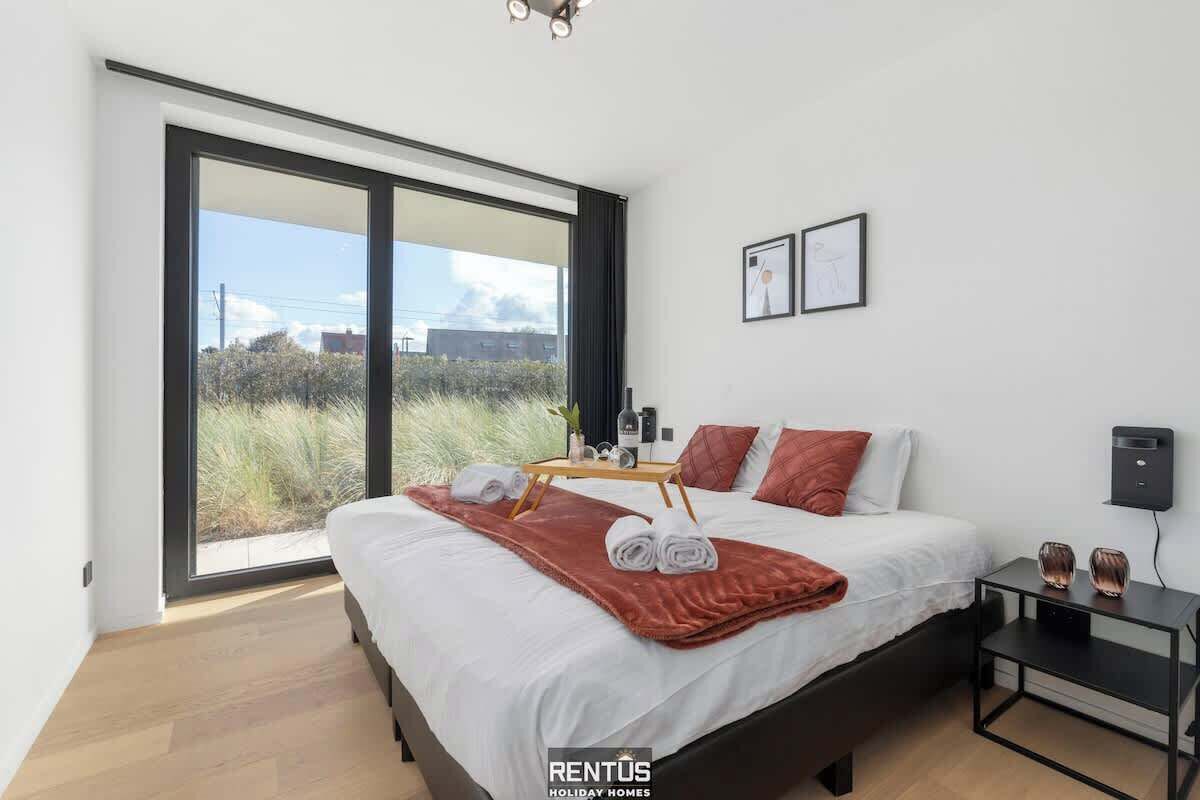 Un dormitorio moderno con un amplio ventanal corredera que da a un balcón, una cama con ropa blanca, una colcha roja y una mesita de noche con dos vasos.